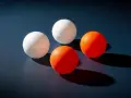 Quatre balles de ping-pong, deux blanches et deux oranges, sont disposées sur une surface sombre. Le volume de chaque balle est bien visible.