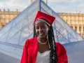 Jeune femme diplômée, souriante, en robe rouge et toque, devant la pyramide du Louvre. Elle prouve qu'on peut avoir le bac avec 9 de moyenne !