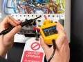 Un électricien vérifie la tension nominale d'un tableau électrique avec un indicateur de tension jaune. Une étiquette "DO NOT OPERATE" est visible.