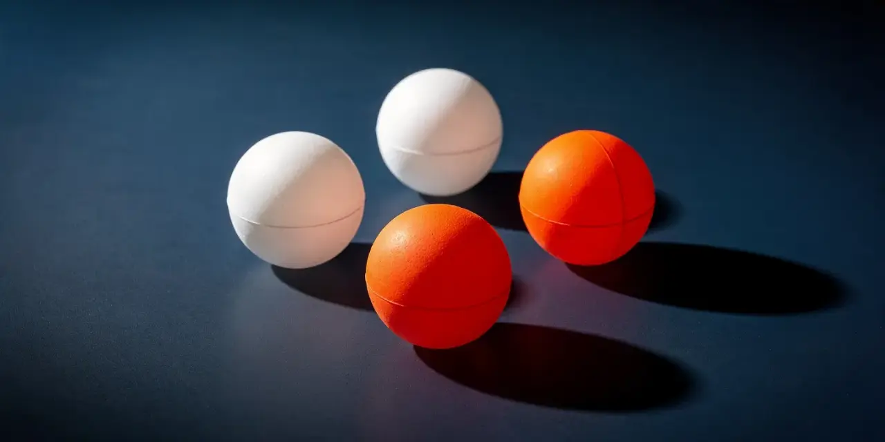 Quatre balles de ping-pong, deux blanches et deux oranges, sont disposées sur une surface sombre. Le volume de chaque balle est bien visible.