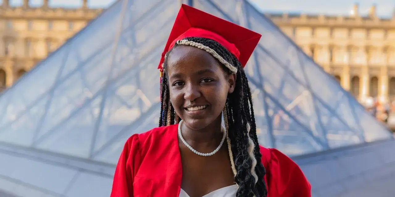 Jeune femme diplômée, souriante, en robe rouge et toque, devant la pyramide du Louvre. Elle prouve qu'on peut avoir le bac avec 9 de moyenne !