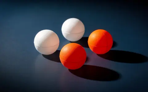 Quatre balles de ping-pong, deux blanches et deux oranges, sont disposées sur une surface sombre. Le volume de chaque balle est bien visible.