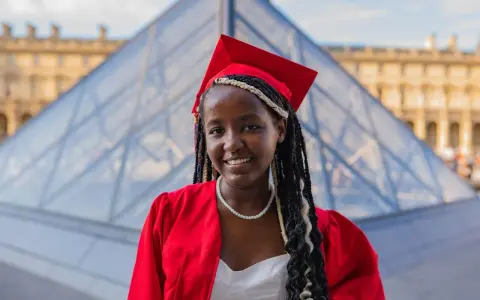 Jeune femme diplômée, souriante, en robe rouge et toque, devant la pyramide du Louvre. Elle prouve qu'on peut avoir le bac avec 9 de moyenne !