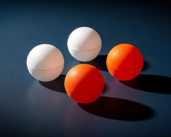 Quatre balles de ping-pong, deux blanches et deux oranges, sont disposées sur une surface sombre. Le volume de chaque balle est bien visible.