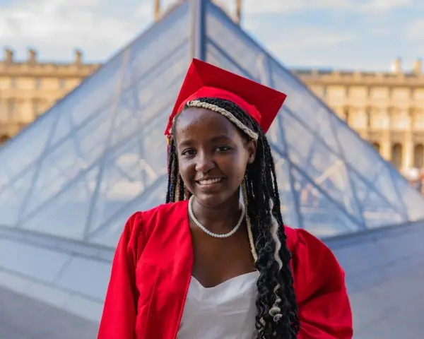 Jeune femme diplômée, souriante, en robe rouge et toque, devant la pyramide du Louvre. Elle prouve qu'on peut avoir le bac avec 9 de moyenne !