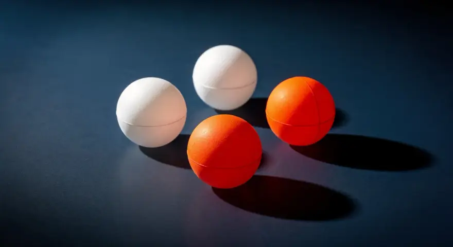 Quatre balles de ping-pong, deux blanches et deux oranges, sont disposées sur une surface sombre. Le volume de chaque balle est bien visible.