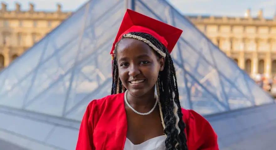Jeune femme diplômée, souriante, en robe rouge et toque, devant la pyramide du Louvre. Elle prouve qu'on peut avoir le bac avec 9 de moyenne !
